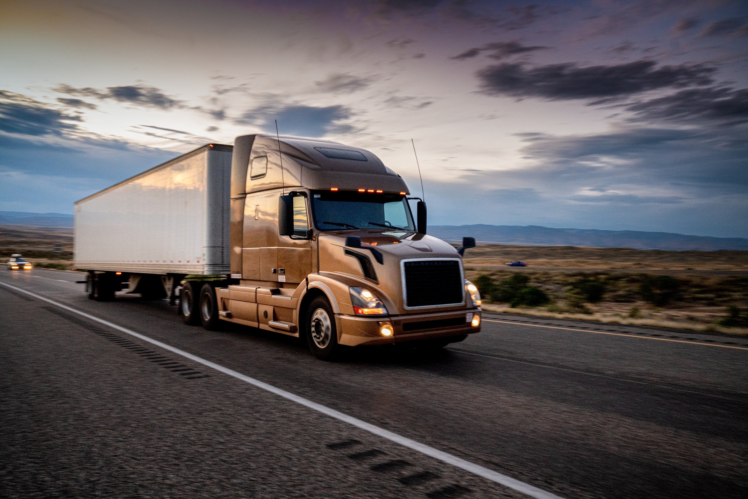 White Semi-Truck Speeding down a four lane highway with a dramatic sunset in the background and headlights on